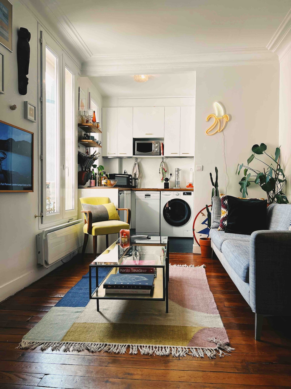 A compact living area features a gray sofa and a glass coffee table centered on a colorful area rug. A small kitchenette is visible in the background, equipped with modern appliances. Decorative plants and wall art add character to the space.