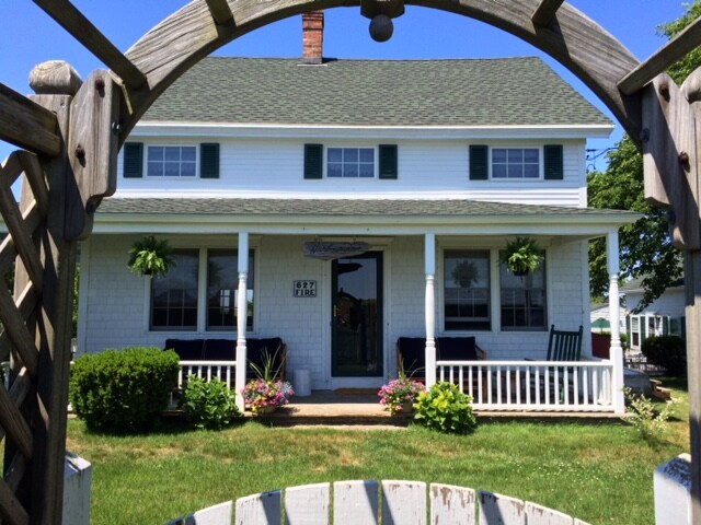 A classic two-story farmhouse is framed by an archway, showcasing a covered front porch with rocking chairs. Green shutters and hanging planters adorn the white exterior. Lush grass and neatly trimmed hedges surround the entrance, inviting guests to relax in the peaceful setting.