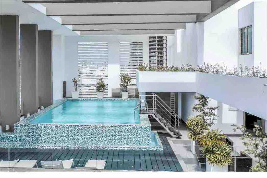 An inviting infinity pool is surrounded by modern architecture and greenery. Sun loungers are positioned nearby for relaxation. Natural light enters through large windows and provides a serene atmosphere. Potted plants enhance the space, contributing to a tranquil environment.