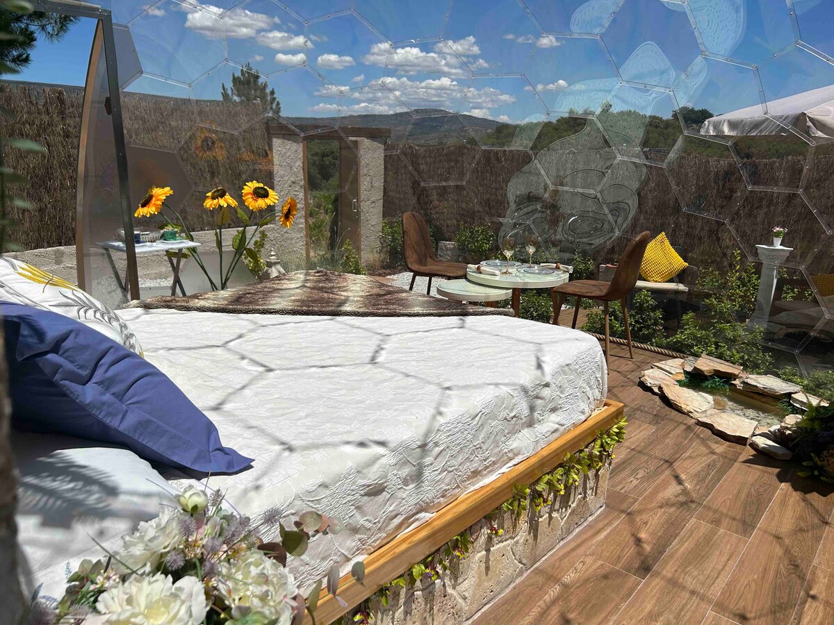A spacious bedroom with a large bed is visible, decorated with neutral bedding and accent pillows. Sunflowers add a pop of color to the interior. Outside, a furnished terrace offers a dining area, surrounded by scenic views and natural foliage.