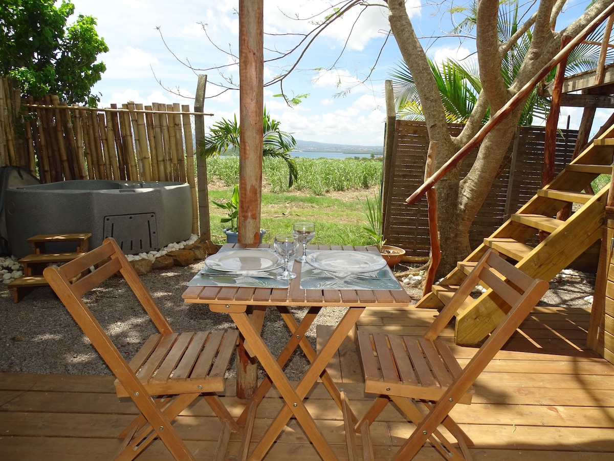 An outdoor dining area features a wooden table set for two, accompanied by light wooden folding chairs. A jacuzzi is located nearby, surrounded by greenery and a wooden fence. Natural light illuminates the space, highlighting the tranquil garden atmosphere.