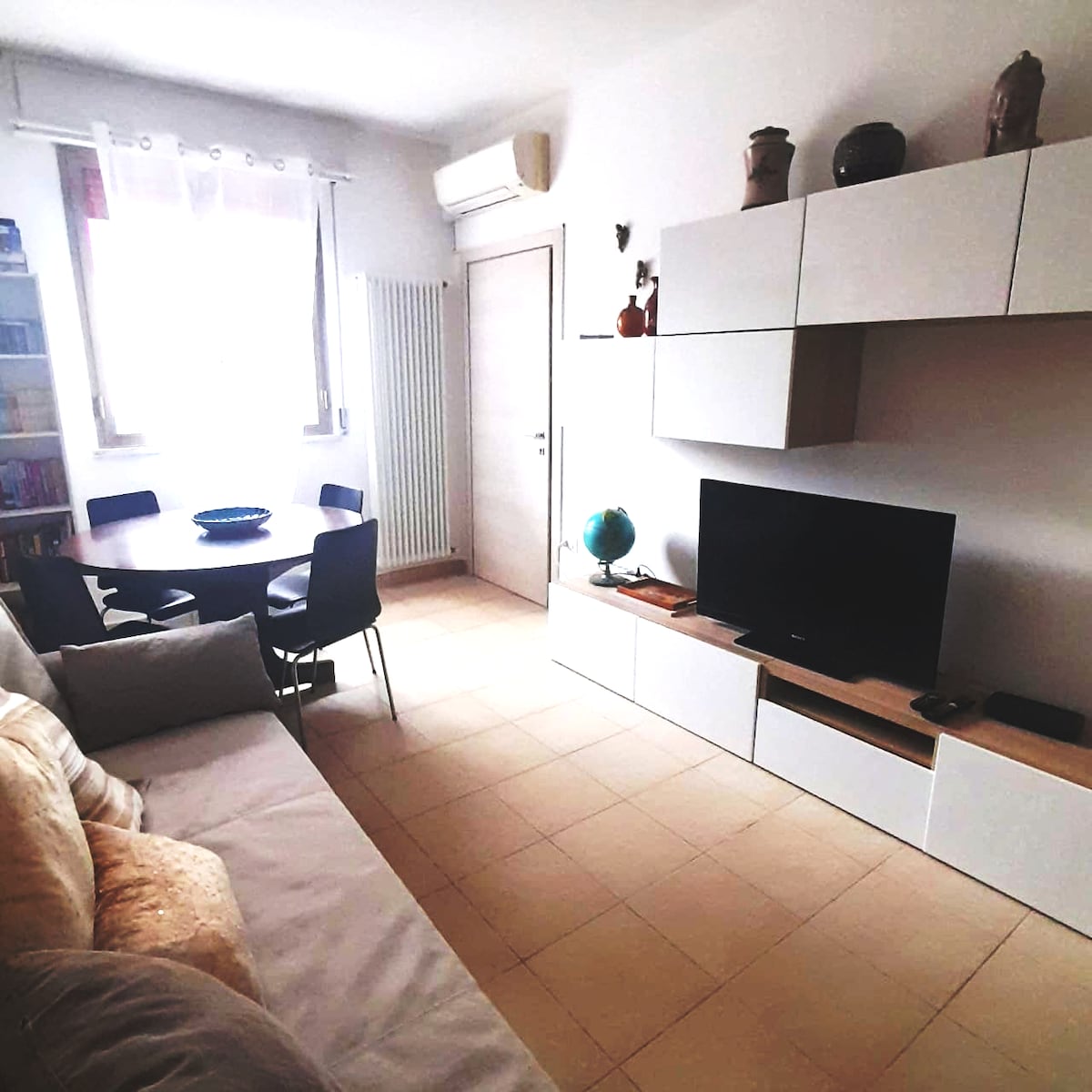 The living area features a comfortable sofa beside a round dining table with four chairs. A television is positioned on a white media unit, next to decorative items on the wall shelves. Natural light fills the space through a large window.