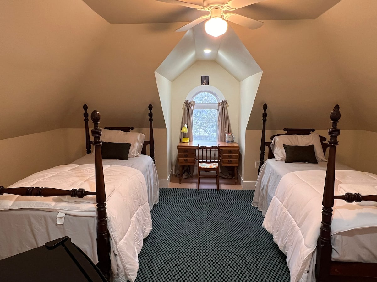 A tranquil room features two twin beds with white bedding, positioned on either side. A wooden desk with a chair is placed under the arched window, which is dressed with light drapes. Soft lighting creates a calm ambiance within the space.