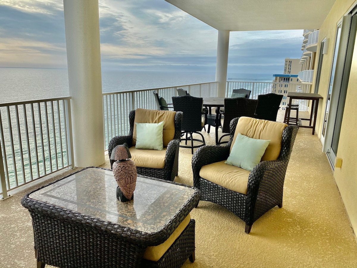 A spacious balcony offers a seating area with wicker chairs and a glass-top coffee table. Light-colored cushions are provided for comfort, while the ocean view extends beyond the railing, creating a serene backdrop for relaxation.