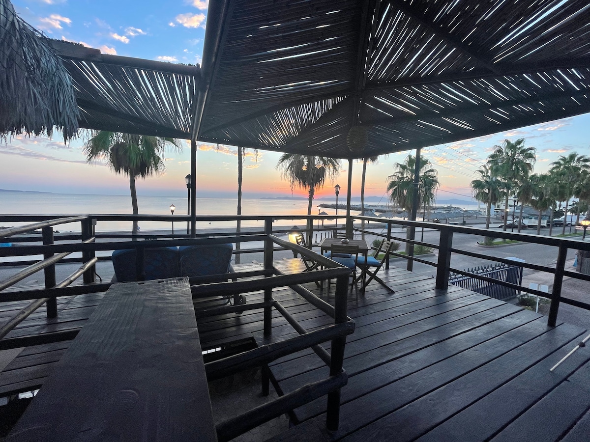 An outdoor seating area features wooden beams and a grass-covered roof, providing shade and a relaxing environment. Comfortable seating is arranged around a dark wooden table, with an unobstructed view of the waterfront, as vibrant sunrise colors reflect off the bay.