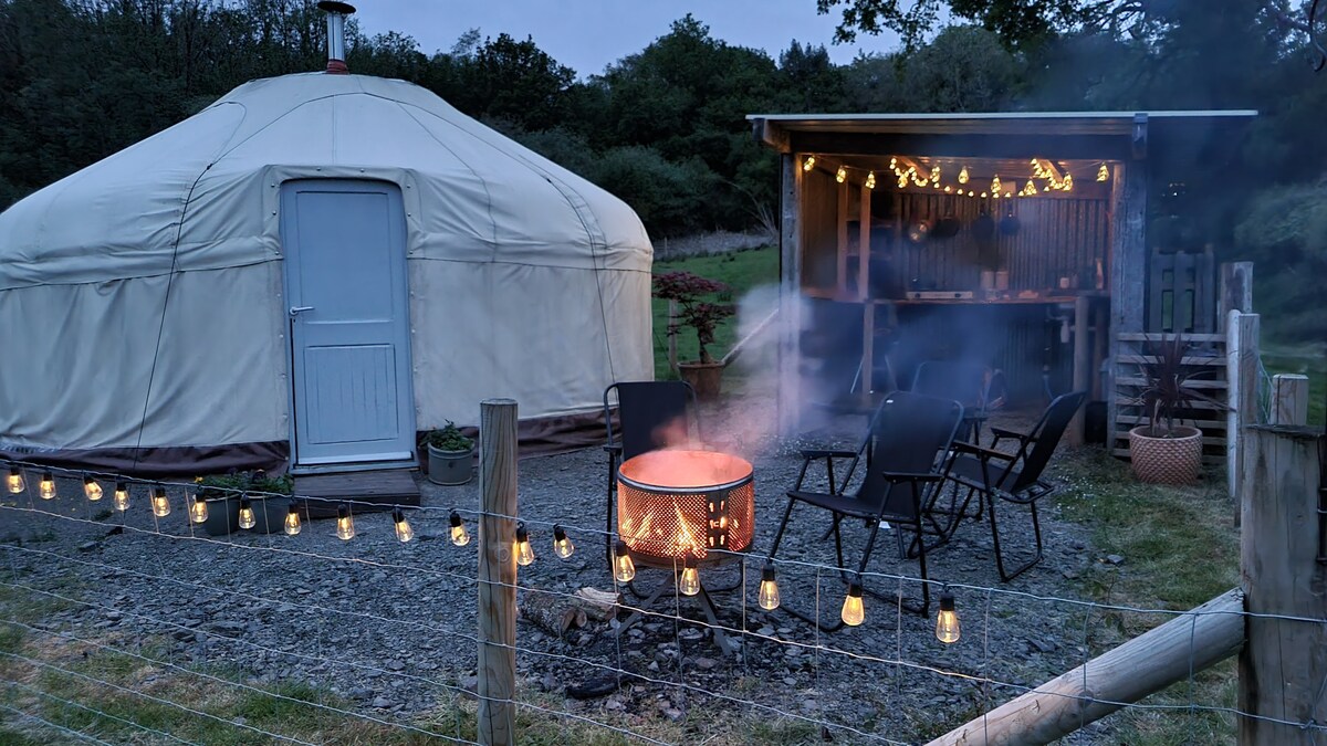 The yurt is complemented by a cozy outdoor area featuring a fire pit surrounded by chairs. Soft lights hang above the outdoor kitchen, creating a warm ambiance. Smoke rises gently from the fire, adding to the serene setting amidst the natural landscape.