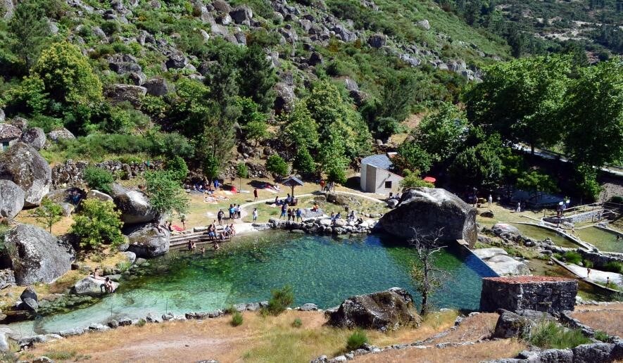 A natural swimming area is surrounded by lush greenery and large boulders. Clear waters reflect the sunlight, while groups of people enjoy the inviting space. Paths and wooden railings can be seen leading into the water, blending harmoniously with the scenic landscape.