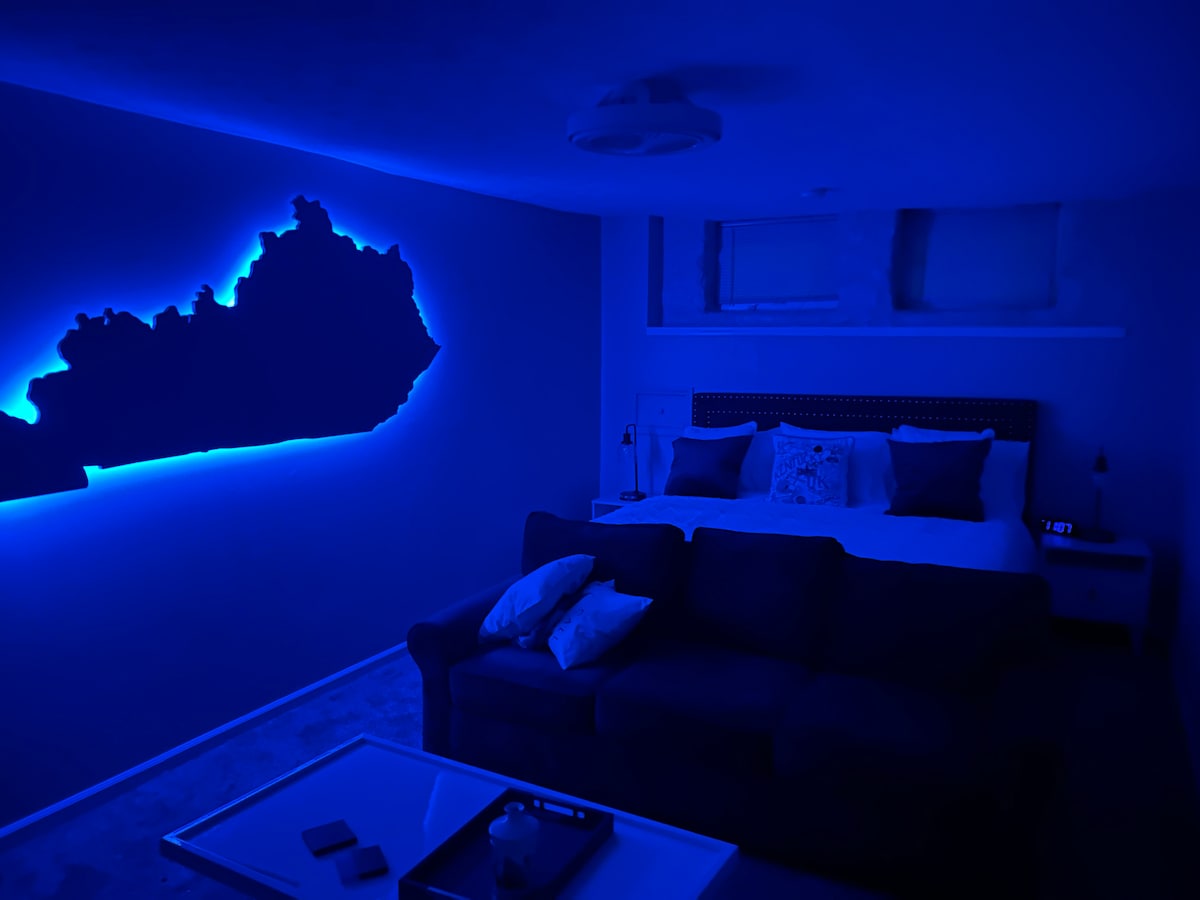 A spacious bedroom features a large king bed with white bedding and decorative pillows. A blue-lit Kentucky silhouette adorns the wall, casting a soft glow. A comfortable blue sofa is positioned nearby, with a coffee table displaying a few items, enhancing the inviting setting.