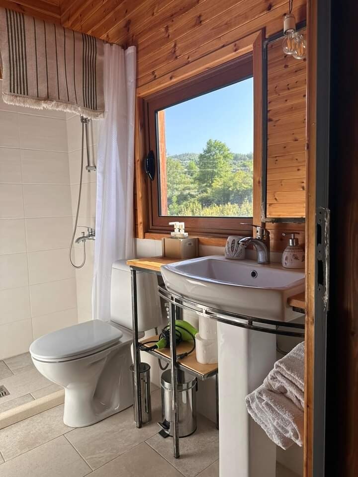 A bright bathroom is presented with natural wood accents. A white ceramic sink is positioned on a metal stand, accompanied by eco-friendly toiletries. A large window allows light to enter, showcasing green hills. A shower area is visible, with simple tiles enhancing the overall cleanliness.