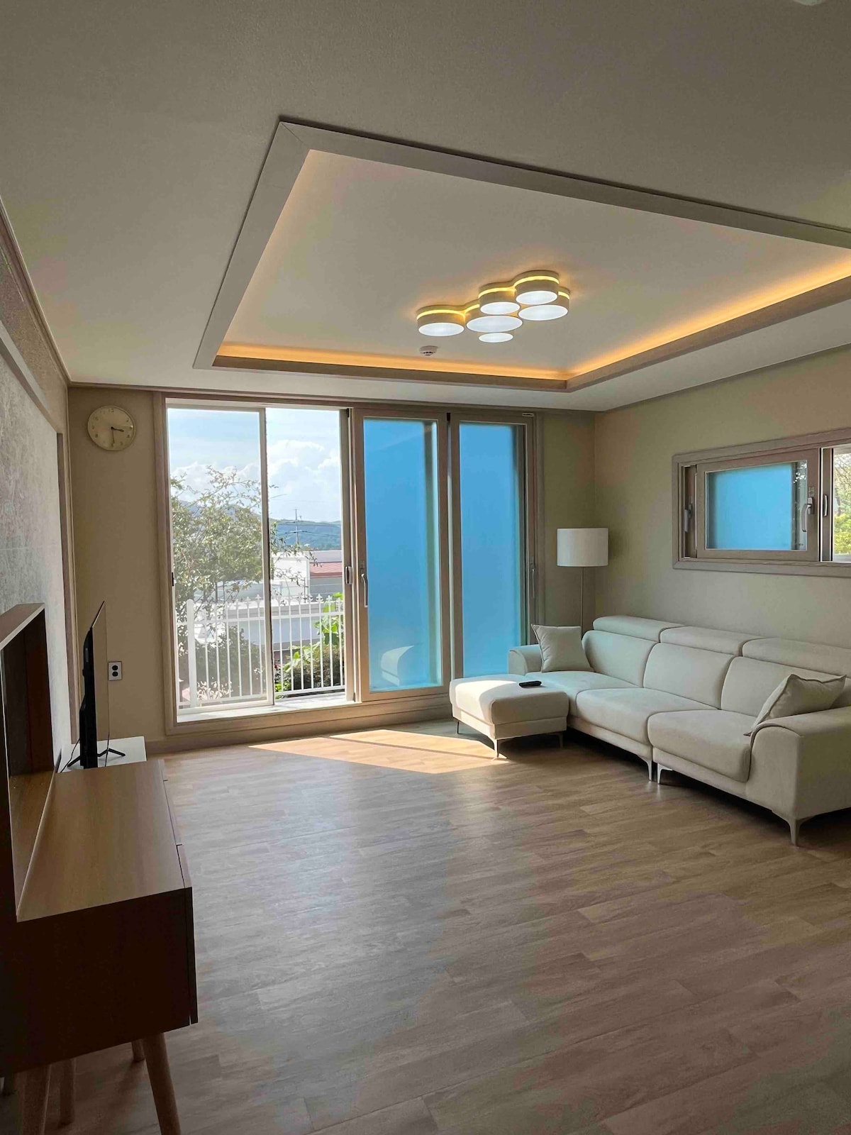 A bright living area features large glass doors allowing natural light to fill the space. A modern white sofa with an accompanying armchair sits against a wall, while a wooden television stand is positioned nearby. Scenic views of the outdoors are visible through the windows.
