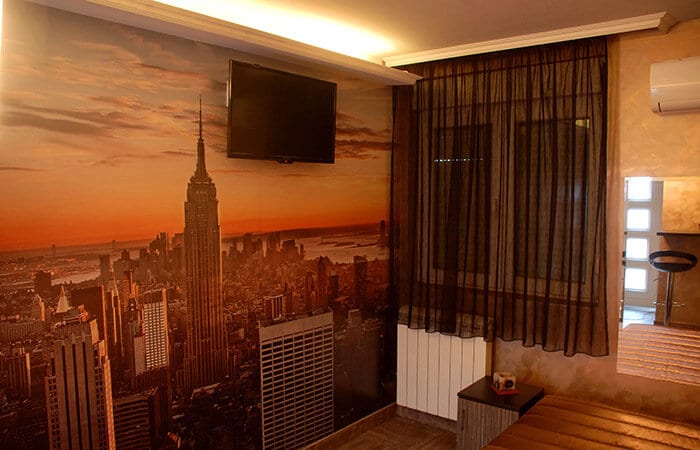 A modern room features a large mural of the New York skyline at sunset. A wall-mounted television is positioned for viewing, and a window is adorned with sheer black curtains, allowing soft light to enter. Minimalist furnishings complement the room's design.