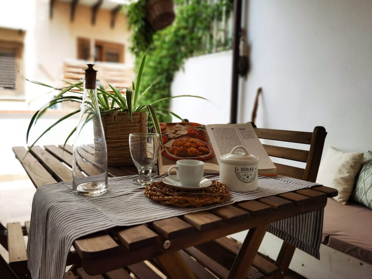 A wooden dining table is set with a glass of water, a small white bowl labeled 'SUGAR,' and a cup placed on a woven placemat. A book and a clay dish with food rest nearby, surrounded by greenery. Soft cushions are visible in the background.