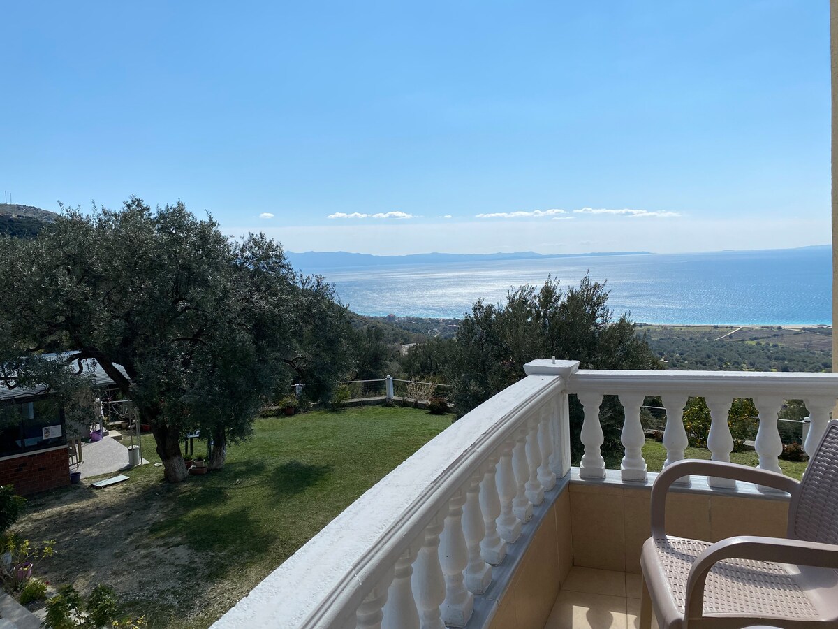 A balcony offers a panoramic view of the sea and distant horizon, framed by olive trees and lush greenery. The peaceful surroundings include a large garden area, with a barbecue space visible below, enhancing the tranquil outdoor experience.