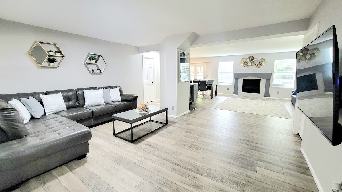 A spacious living area features a modern sectional sofa with decorative pillows, complemented by a sleek coffee table. Wall-mounted decor and a large flat-screen TV enhance the room. Large windows allow natural light to fill the space, leading into an adjacent dining area.