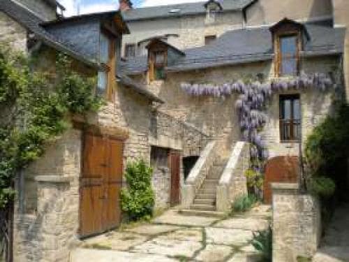 A charming stone building is depicted with wooden doors and windows, characterized by rustic architecture. Wisteria blooms cascade over the structure, enhancing the natural beauty of the space. A set of stone steps leads to upper levels, framed by greenery on either side.