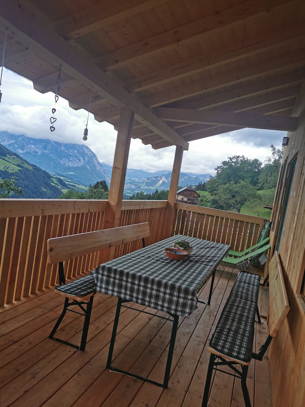 An outdoor balcony area is presented, featuring a wooden ceiling and railing. A checkered tablecloth covers a rectangular table, surrounded by benches. A green chair complements the rustic setting, with scenic mountain views visible in the background beneath a partly cloudy sky.