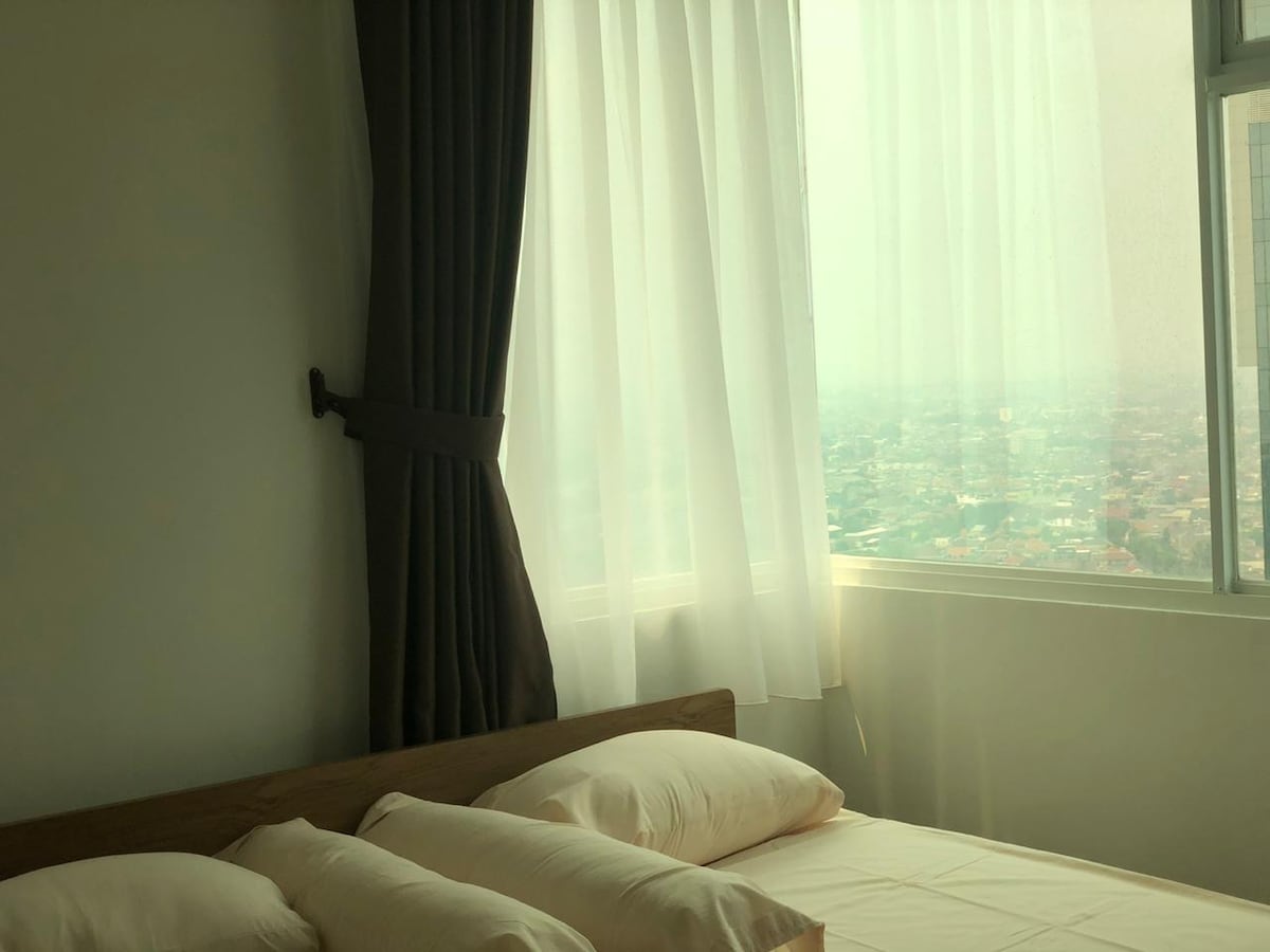 A light-filled bedroom features a spacious bed with neutral-toned linens. Soft, sheer curtains frame a large window, offering a view of the cityscape. The overall ambiance conveys a serene and calming space.