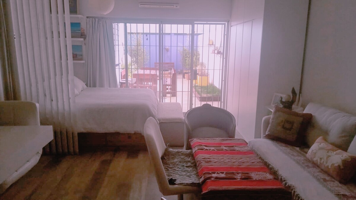 A bright and open living space features a spacious bed in the background and a comfortable seating area in the foreground. Soft textiles add warmth to the room, while large glass doors open to a private patio, allowing natural light to fill the interior.