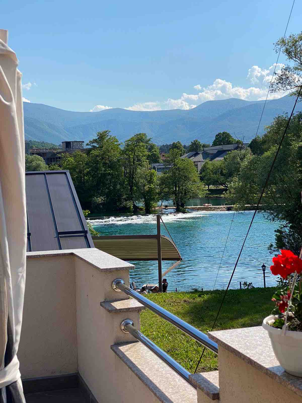 A serene view of the river Una is presented, framed by lush greenery and distant mountains. The image showcases a balcony railing and a bright red flower pot, adding a splash of color. Sunlight reflects on the water, enhancing the peaceful atmosphere.