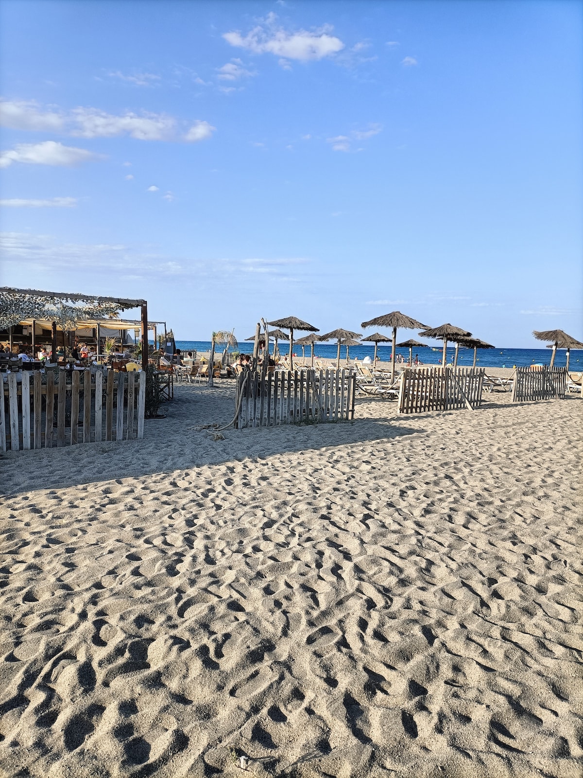 A sandy beach scene is visible, featuring thatched umbrellas providing shade for lounging areas. Wooden fences are positioned within the sand, delineating seating options. The calm blue sea stretches in the background under a clear sky with a few scattered clouds.