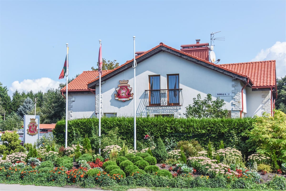 A cozy building with a red-tiled roof showcases elegant architecture. Lush greenery surrounds the property, featuring well-maintained flower beds and shrubs. National flags are displayed in front, adding a touch of local character to the inviting entrance.
