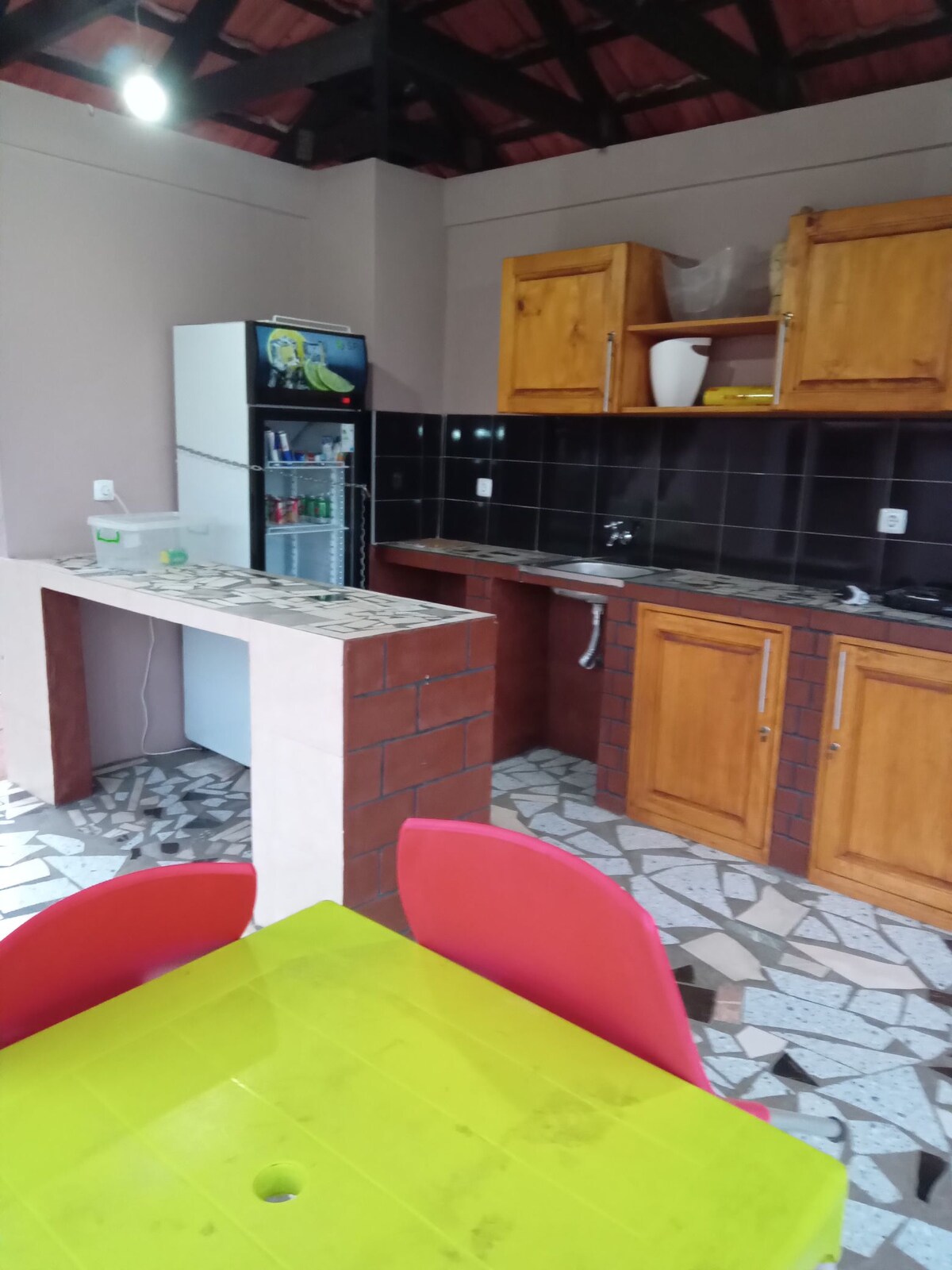 A functional kitchen area features wooden cabinetry with a dark tiled backsplash. The space includes a refrigerator, a sink, and open shelving. A dining table with bright red chairs is positioned in the foreground, complemented by a vibrant yellow tabletop.
