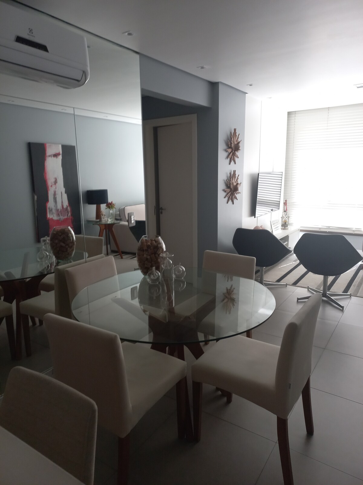 A modern dining area features a round glass table surrounded by four light-colored chairs. A mirrored wall enhances the sense of space. In the background, a stylish seating area with a black chair and a small lamp is visible, complemented by bright natural light coming from the window.