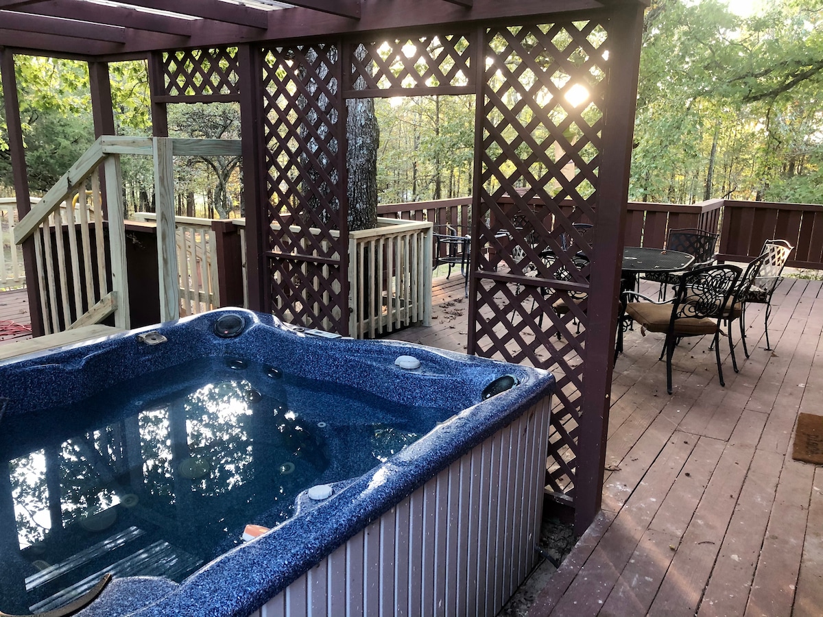 A private hot tub is situated within a wooden pergola, surrounded by a lattice design. A spacious deck area features a dining set with chairs and a table, illuminated by soft natural light filtering through the trees.