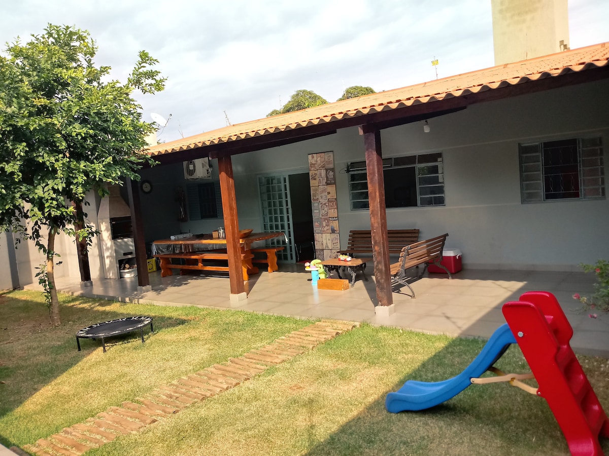 An outdoor area features a covered patio with wooden furniture and a barbecue setup. A trampoline can be seen on the grassy lawn, along with a small slide for children. The space is surrounded by greenery, creating a relaxing environment for families and friends.