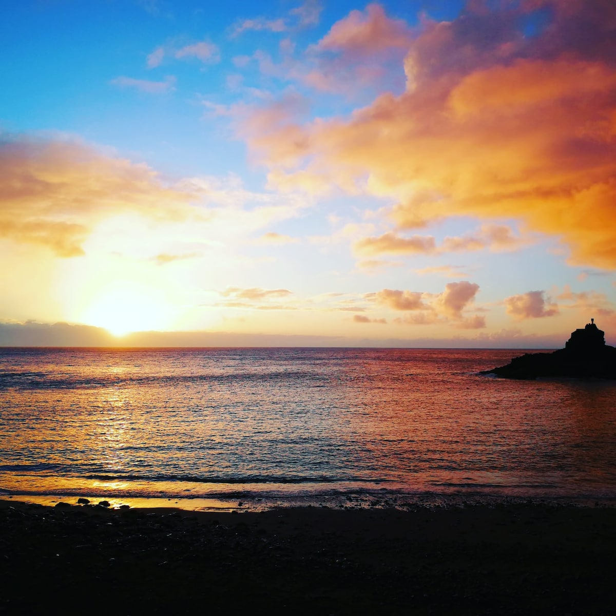 A tranquil seascape captures the sun setting over the horizon, casting warm hues of orange and pink across the sky. The calm waters reflect these colors, while a rocky formation is visible in the distance, creating a serene atmosphere.