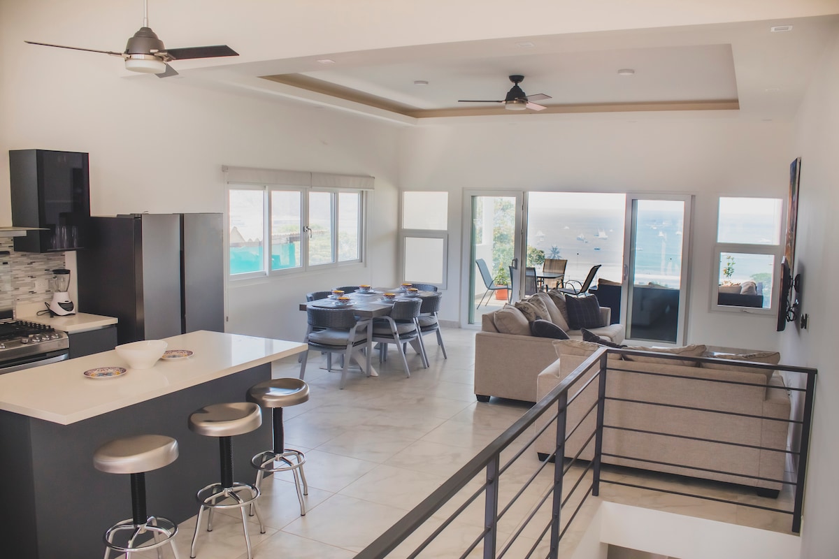 An open-concept living space features a modern kitchen with stainless steel appliances and a breakfast bar. Comfortable seating is arranged for socializing near large windows that offer views of the ocean. Natural light fills the area, enhancing the spacious layout.
