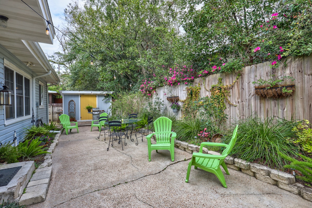 A charming courtyard is surrounded by lush foliage and colorful flowers. Several green chairs are arranged around a small black table, inviting relaxation. A decorative fence lined with hanging planters adds visual interest, while a shaded area is created by overhanging trees.