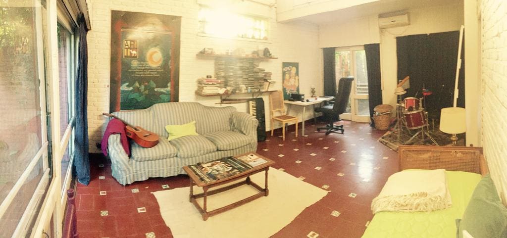 A spacious living area is presented, featuring a striped couch and a coffee table surrounded by decorative items. A desk area with a computer is visible, alongside a drum set. The space is illuminated by natural light from large windows, enhancing its openness.