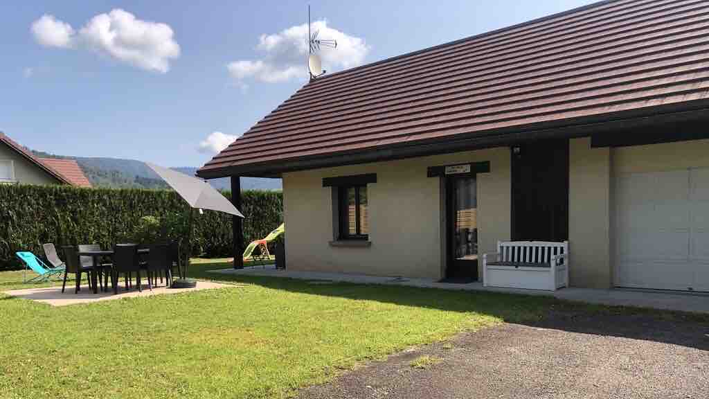 A charming house surrounded by a lush green lawn features a shaded terrace with outdoor dining furniture. A vibrant yellow slide is present, along with a sun umbrella. The entrance is framed by large windows, providing natural light to the interior.