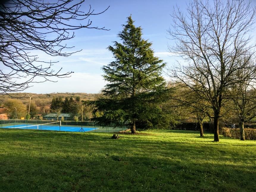 A serene outdoor space features a vibrant green lawn, framed by tall trees. In the background, a tennis court is visible, highlighting blue asphalt against the natural landscape. The area is surrounded by lush greenery, creating a peaceful atmosphere.
