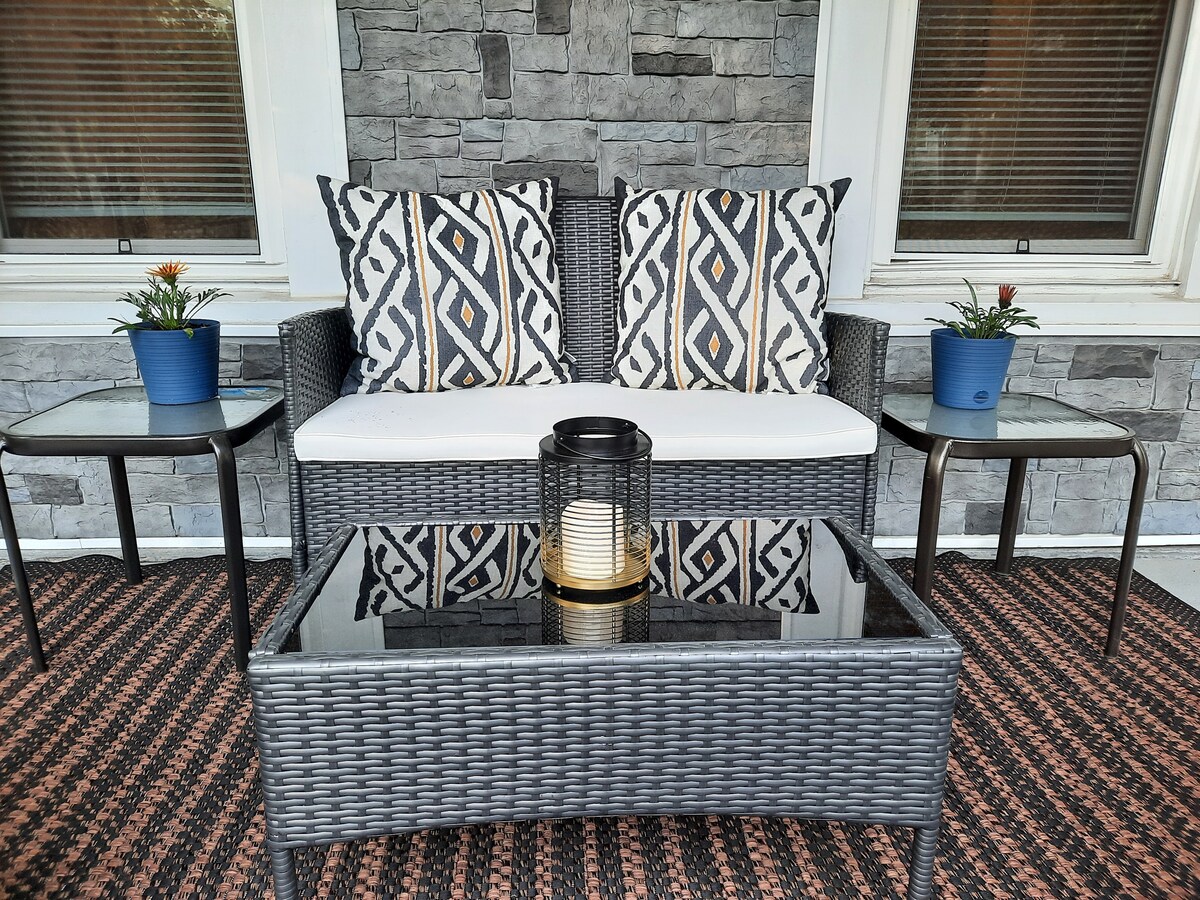 A welcoming outdoor seating area features a wicker sofa adorned with patterned throw pillows. Two side tables flank the sofa, with potted plants placed on each. A glass-top coffee table holds a lantern, adding a functional and decorative touch to the space.