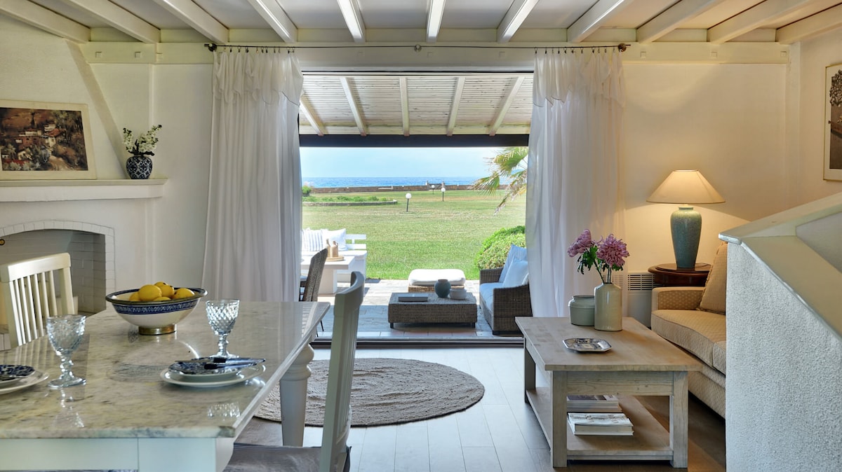 A light-filled living area features a comfortable sofa and a coffee table with decorative items. Large, open glass doors reveal scenic views of the lush green lawn and Aegean sea beyond, seamlessly connecting indoor and outdoor spaces.