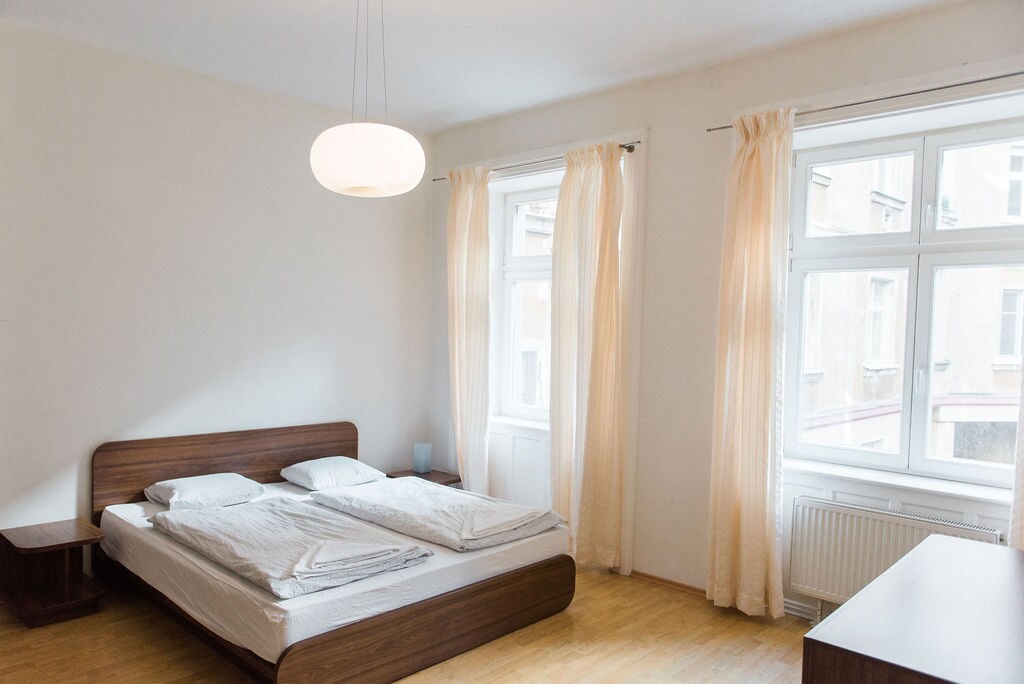 A spacious bedroom is revealed with two beds arranged side by side, topped with crisp white linens. Natural light filters through two large windows adorned with sheer, light-colored curtains, creating a bright atmosphere. A minimalist bedside table is positioned next to the beds, enhancing the room's simplicity.
