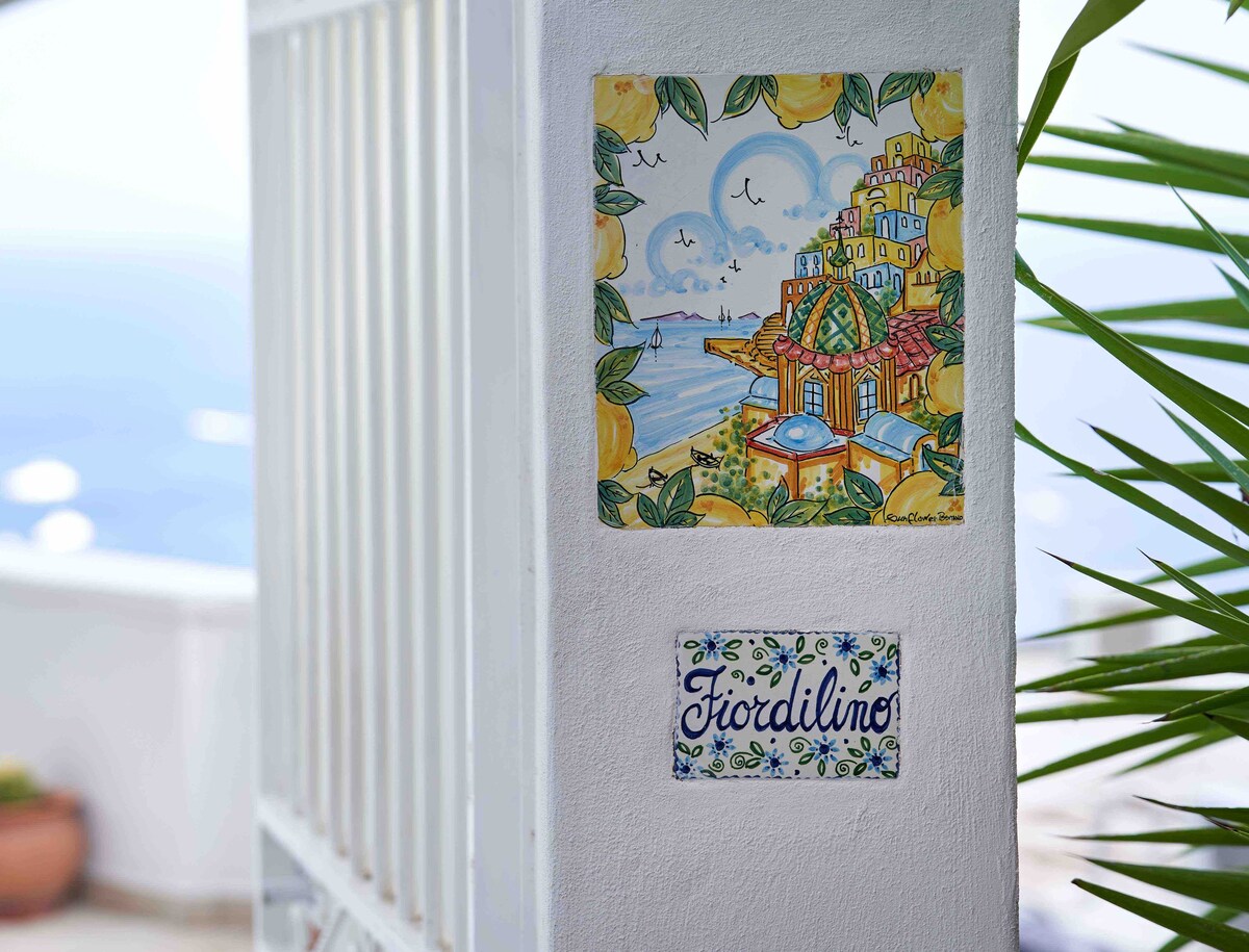A vibrant wall displays two decorative tiles. One features a colorful illustration of coastal scenery with a lemon motif, while the other shows the name "Fiordilino" in an artistic design. Green foliage is visible beside the wall, adding to the pleasant ambiance.