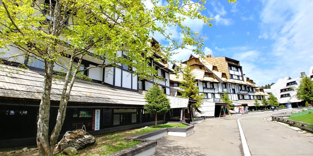 A modern building complex is surrounded by greenery, featuring traditional architectural elements. A tree with fresh green leaves provides shade, while a pathway leads towards the entrance. Children’s playground equipment is visible, contributing to a family-friendly environment.