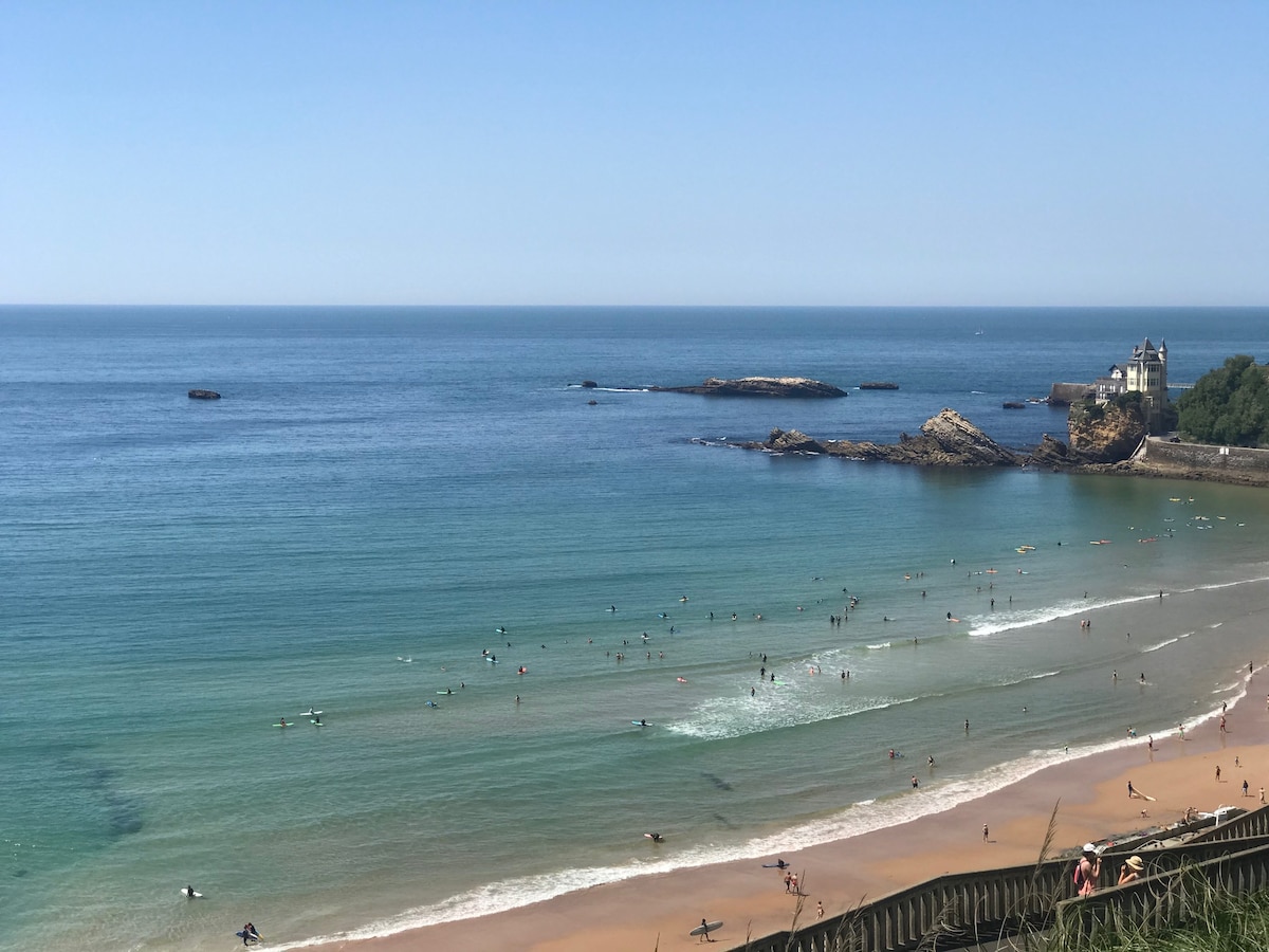 A wide view of a sandy beach is depicted, with gentle waves lapping at the shore. Surfers and beachgoers are scattered across the water and sand, enjoying a sunny day. A rocky outcrop can be seen in the distance, alongside a charming coastal building.
