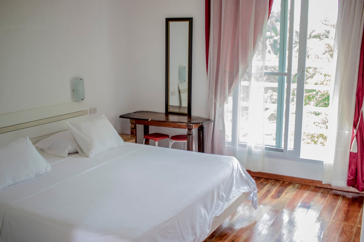 A well-lit bedroom features a spacious bed with crisp white linens, complemented by decorative pillows. A wooden table with two stools adds functionality, while a mirror reflects natural light. Sheer curtains frame a window, allowing views of the greenery outside.