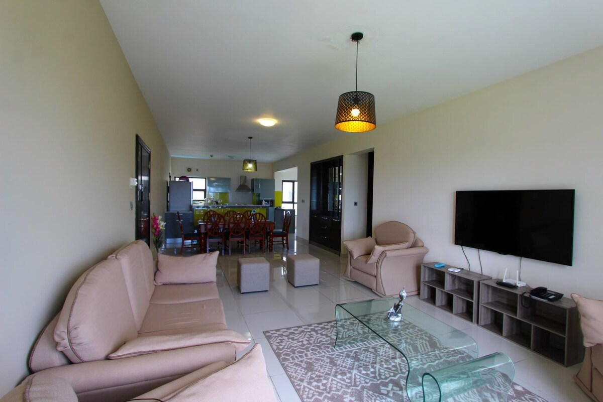 A light-filled living area features soft beige couches and a glass coffee table on a textured area rug. A mounted flat-screen television is positioned on a low wooden console. A dining area is visible in the background, complemented by modern light fixtures.