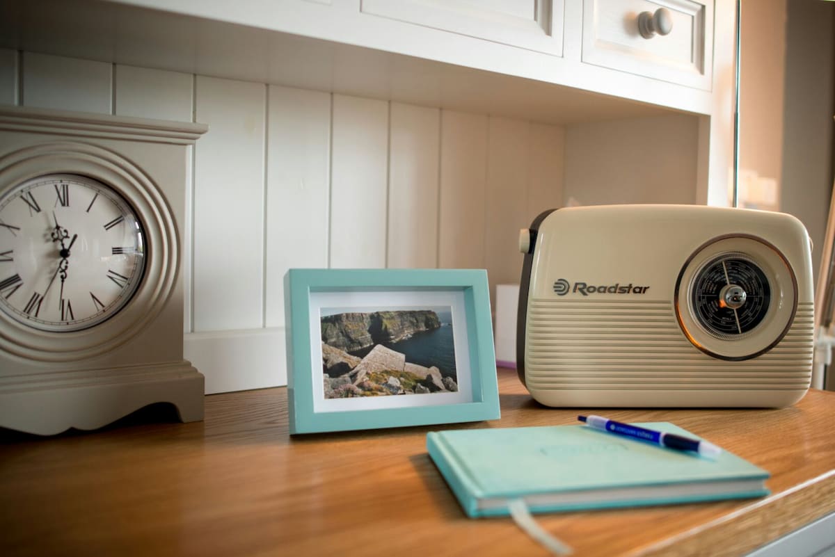 A wooden surface features a vintage clock and a retro radio alongside a light blue picture frame displaying a scenic image. A notepad and pen are placed nearby, suggesting a cozy workspace atmosphere.