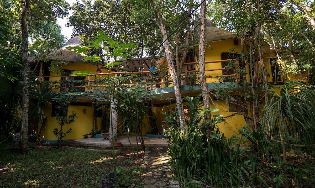 A vibrant yellow bungalow is nestled among lush greenery, showcasing a wraparound balcony surrounded by trees. The structure features a thatched roof, blending harmoniously with the tropical environment. Pathways meander through the garden, adding to the serene ambiance of the space.