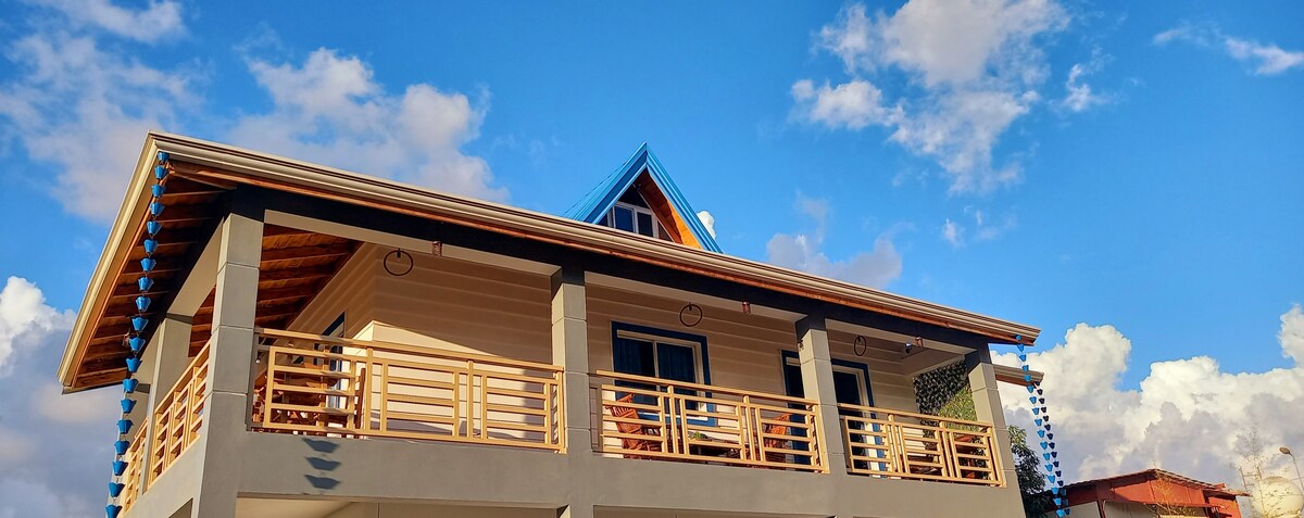 The two-story building features a gabled roof with bright blue accents. A spacious balcony is adorned with wooden railings, while large windows provide natural light. The sky above is dotted with fluffy clouds, enhancing the tranquil setting.