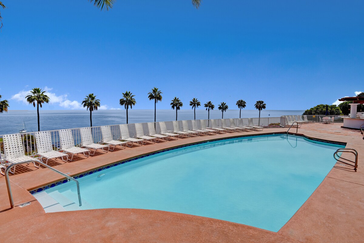 A sparkling outdoor pool is surrounded by abundant lounge chairs, offering a relaxed area for sunbathing. Lush palm trees overlook the tranquil ocean, creating a serene atmosphere for guests. The clear blue sky complements the vibrant scenery.