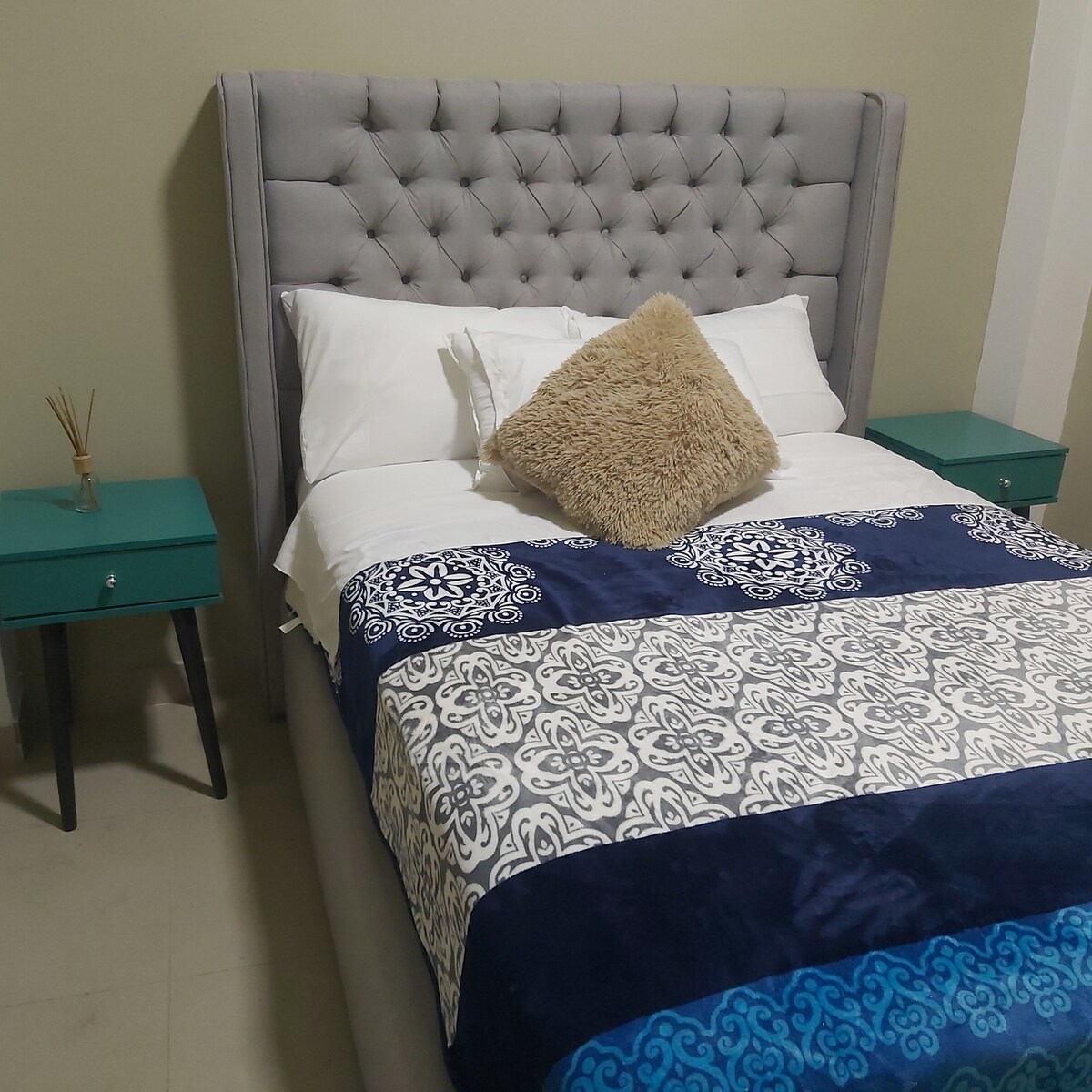 A spacious bed is featured with a tufted grey headboard, adorned with a decorative pillow. The bedding has a mix of patterns in navy and white tones. Two small teal nightstands are positioned on either side, complementing the calming room ambiance.