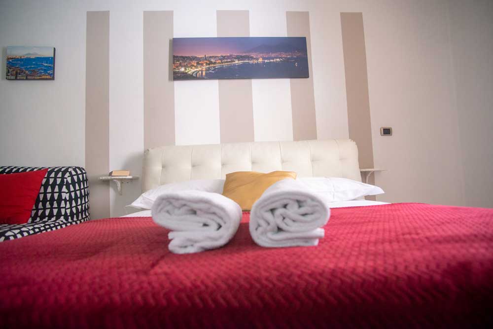 A neatly arranged bed is seen at the center of the image, featuring a soft white headboard and a red bedspread. Two freshly laundered towels are positioned at the foot of the bed. A scenic wall art piece adds visual interest above the headboard, while striped wallpaper provides a subtle backdrop.