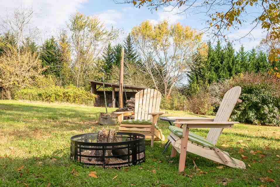 A spacious backyard is shown, featuring two wooden chairs positioned around a fire pit. A pile of firewood and trees are visible in the background, alongside an open area of grass, ensuring a tranquil outdoor setting.